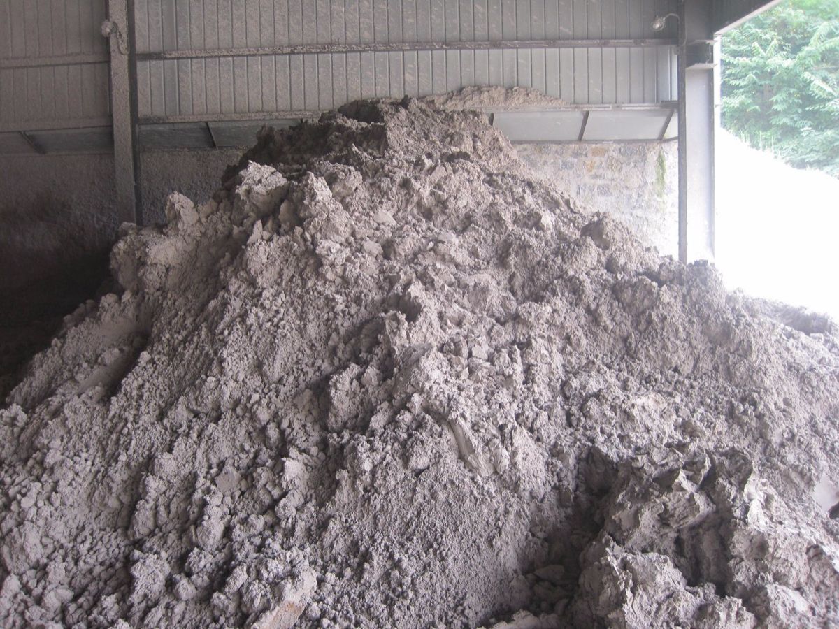 A heap of raw grey clay, in lumps and powder, piled several metres high inside a steel-framed shed.