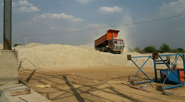 A tipper truck with its bed raised, standing on a pale earth stockpile, dust hanging in the air.