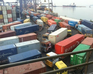 A port yard with containers stacked several high and on trailers, gantry cranes behind, a ship in open water beyond.